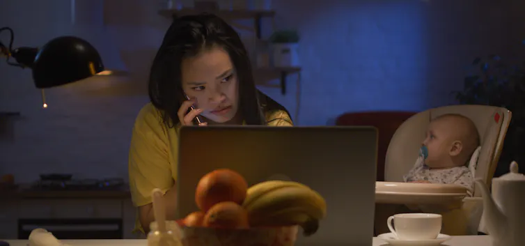 A woman working on a laptop with a baby beside her in a high chair
