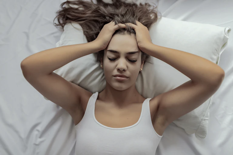 A young woman, sick and lying in bed, has her hands placed on the sides of her head.