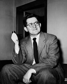 A dark-haired man in a suit and tie, wearing glasses and sitting down, looking happy.