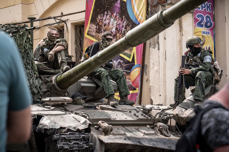 Several men, clad in army fatigues, sit atop an army tank. Behind them, a decorative banner hangs from a building.