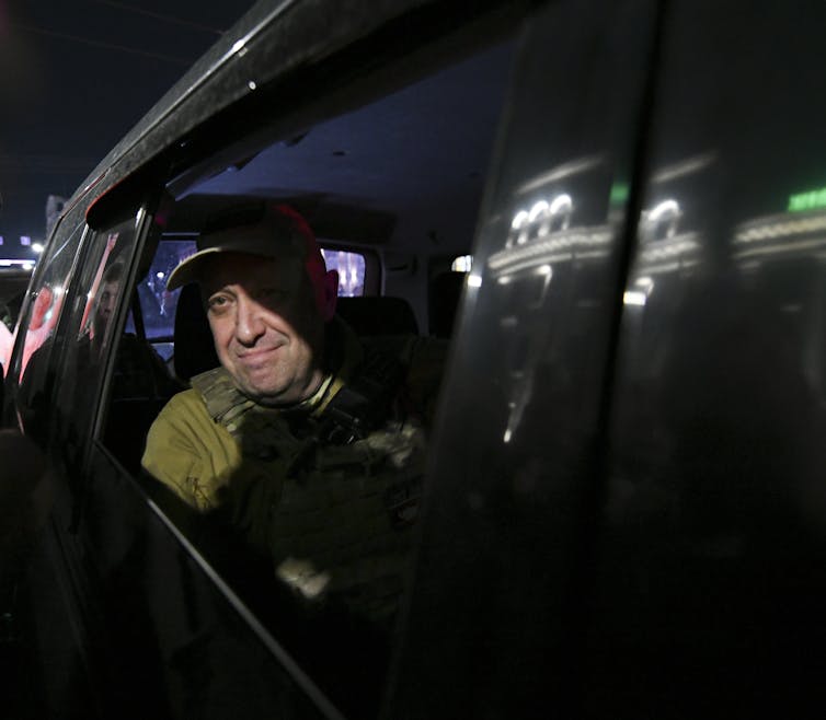 A man wearing a green army cap and uniform looks out an open car window at night.