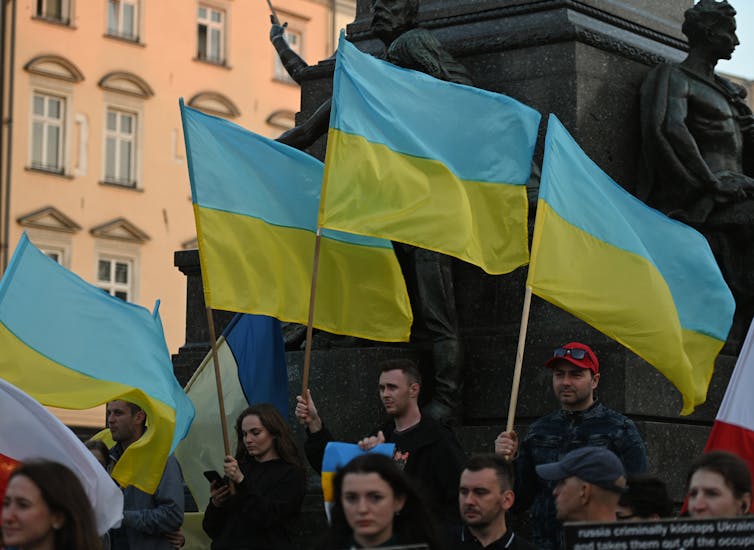 Men and women stand in a street holding the blue and yellow Ukrainian flag as it blows in the wind.