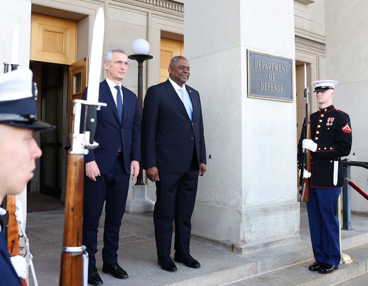 Two men, wearing suits, stand with their hands to the sides. To their left, a man in a US marine uniform stands guard holding a rifle that is pointed up.