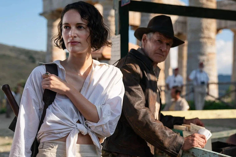 A woman in a white shirt holds a rucksack handle and looks off camera. A man behind her slightly off focus with a fedora hat.