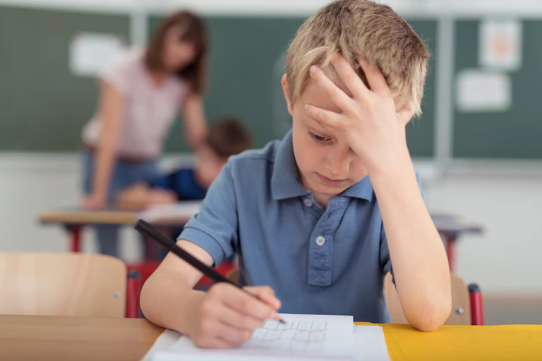 boy sits at school desk with hand on forehead