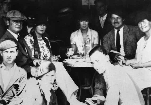 Un grupo de gente se sienta alrededor de una mesa en un bar en los años 20.