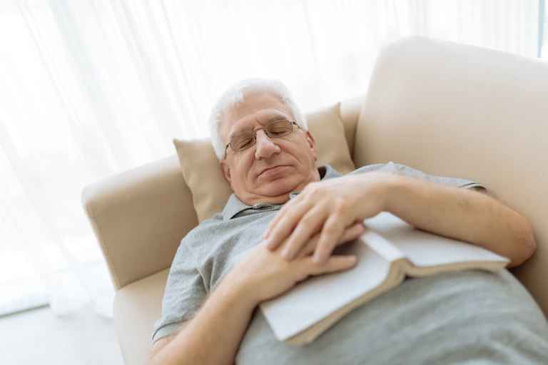 An elderly man napping on the couch with a book on his chest.