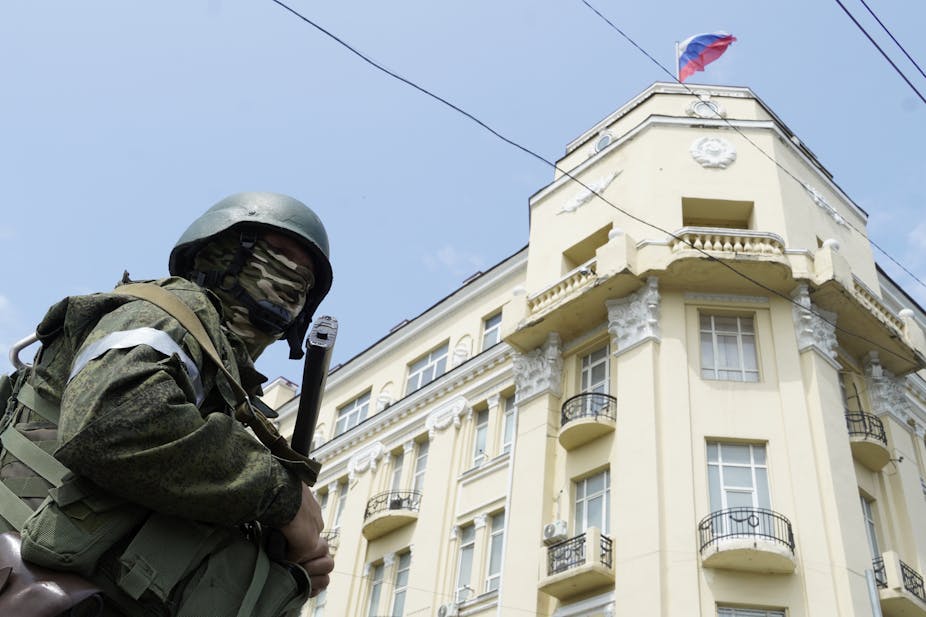 Militaire au visage masqué armé d'un fusil, devant un bâtiment sur lequel flotte un drapeau russe