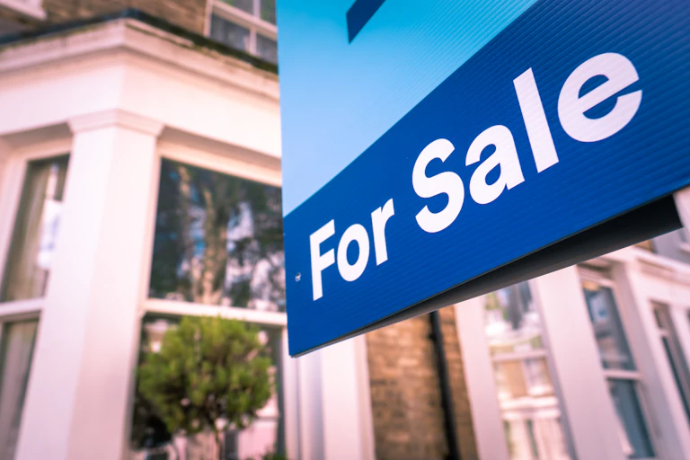 Blue For Sale sign in front of the bay window of a home with the reflection of a green tree.