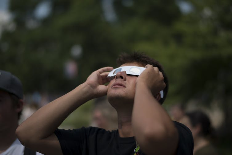 A man looking up at the sky, wearing gray paper eclipse glasses.