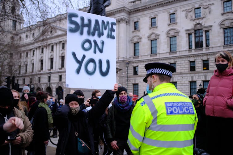 A Met police officer stands facing a protester who holds a large sign reading 'shame on you'