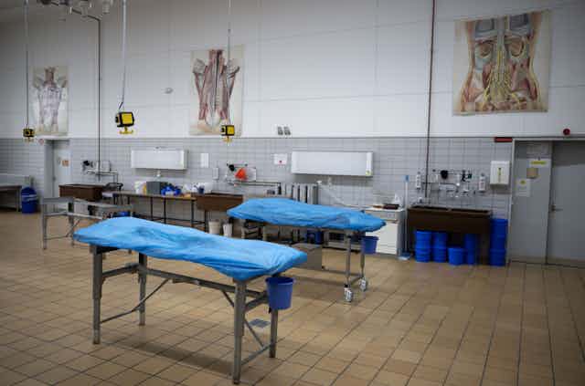 Two metal tables in a large, tile-covered room have blue covers concealing bodies.