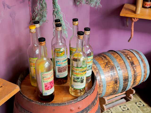 Eight glass bottles filled with liquor sit atop a wooden barrel.