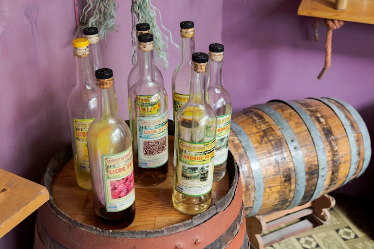 Eight glass bottles filled with liquor sit atop a wooden barrel.