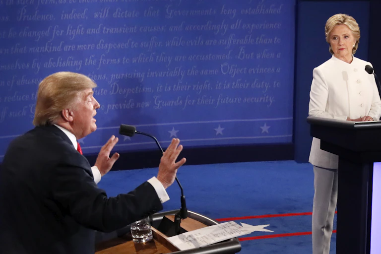 A man in a black suit speaks from a lectern and gestures with both hands up, palms facing out as a woman in a white suit stands behind a lectern looking at him.