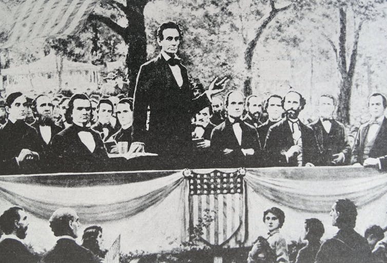 A tall, suited man stands with his right hand in a tented position on a table beside him and his left hand raised, palm facing up. Behind him, a crowd of men, also wearing suits, sit and look in his direction.
