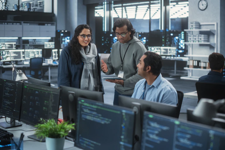 Three people in casual work attire have a conversation between two rows of computer desks