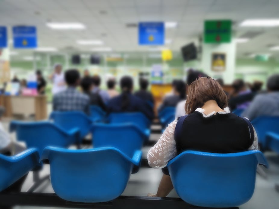 A woman sits alone in a row of blue plastic chairs in a clinical setting