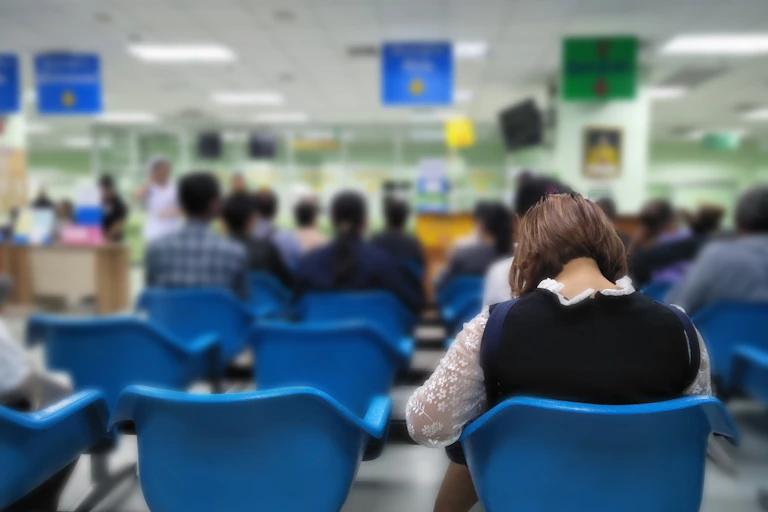 A woman sits alone in a row of blue plastic chairs in a clinical setting