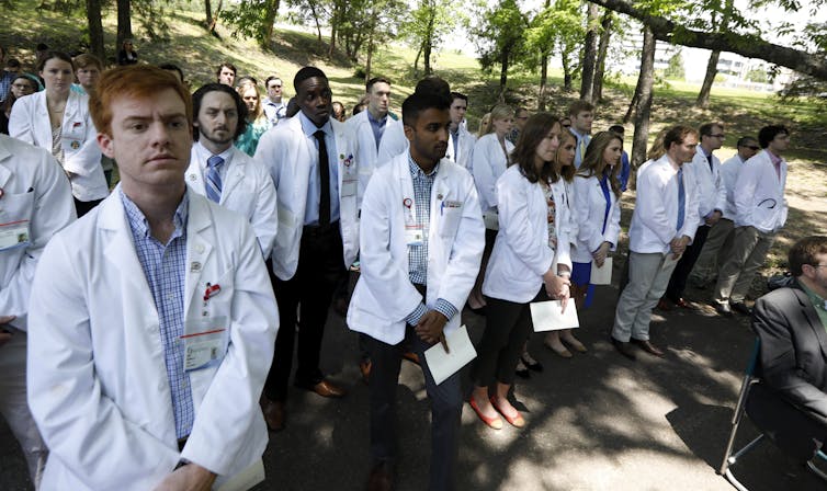Rows of young people in white medical coats stand respectfully outside at a ceremony.