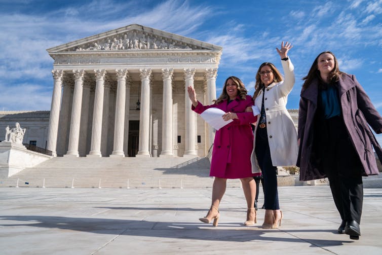 Three women in coats wave as they walk away from a huge building with tall pillars.