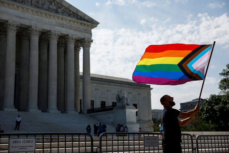 A man holding a rainbow-striped flag waves it in front of an ornate building with pillars.