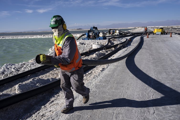 A worker carries a large hose along the edge of a turquoise lithium pond. The worker is wearing a facemask against the dust and reflective gear.