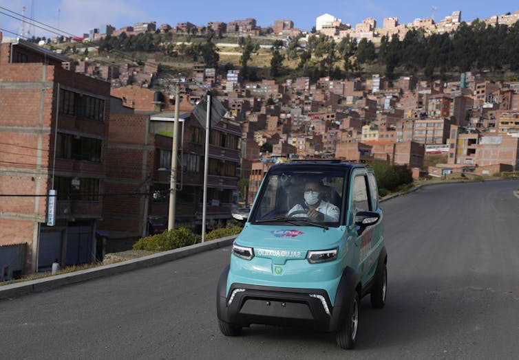 A tiny car big enough for one person, with no passenger seats, drives down a street of brick buildings. Quantum Motors, its maker, is based in Bolivia.