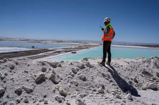 A man with a walkie talk and reflective construction zone vest stands on a pile of lithium that looks like white sand overlooking large turquoise ponds stretching into the distance. This is Chile's lithium mining region, where China has been investing in resources essential for clean energy.