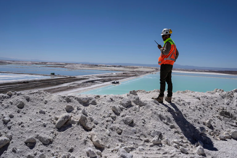 A man with a walkie talk and reflective construction zone vest stands on a pile of lithium that looks like white sand overlooking large turquoise ponds stretching into the distance. This is Chile's lithium mining region, where China has been investing in resources essential for clean energy.