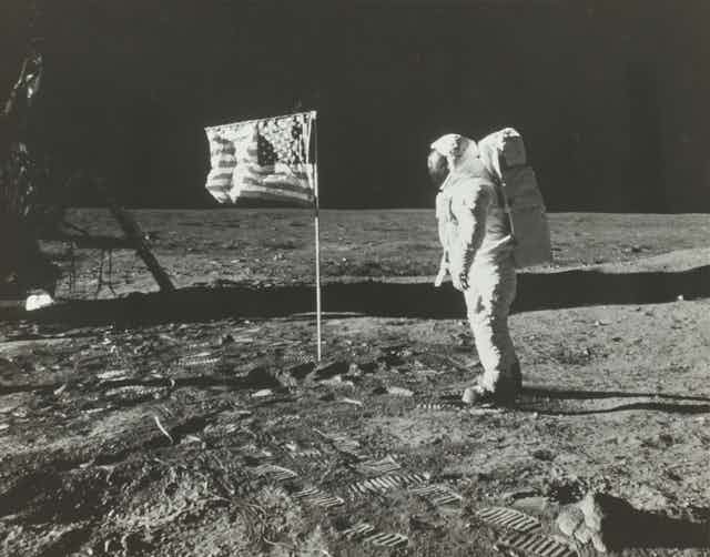 Standing on the Moon's dusty surface, an astronaut pauses to look at the U.S. flag.