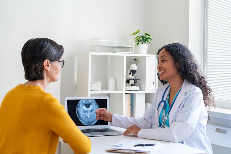 A smiling health care worker in a white lab coat sits across from a patient and points at an x-ray scan on a screen.