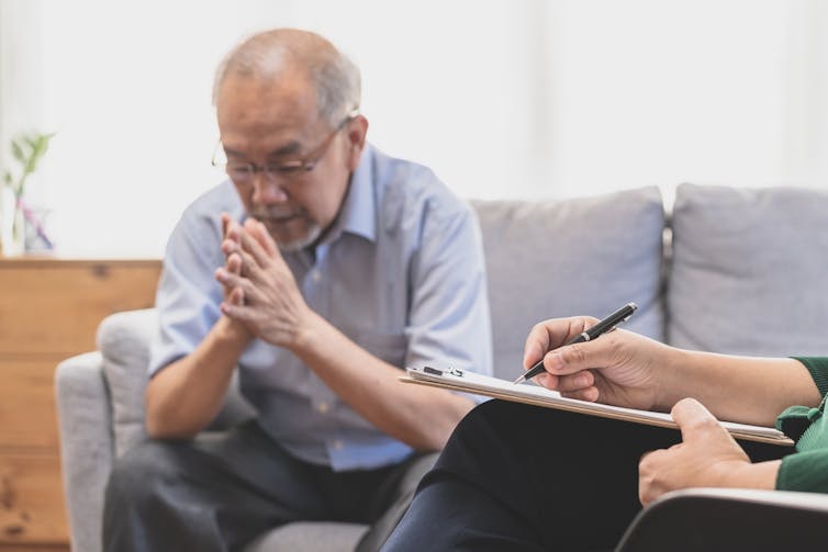 Older man, sitting on sofa, palms together, with therapist taking notes