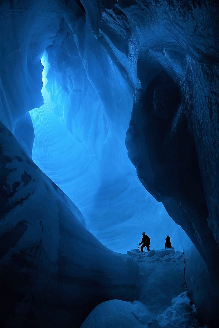 Two people stand inside an ice cave with light coming from a large hole above.