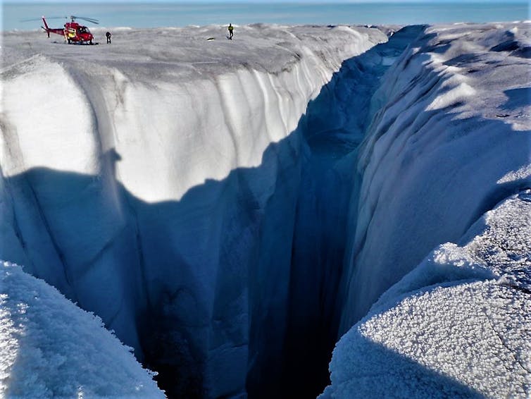 A helicopter sitting on the ice sheet looks tiny next to the gaping moulin, where a meltwater stream pours into the ice sheet.