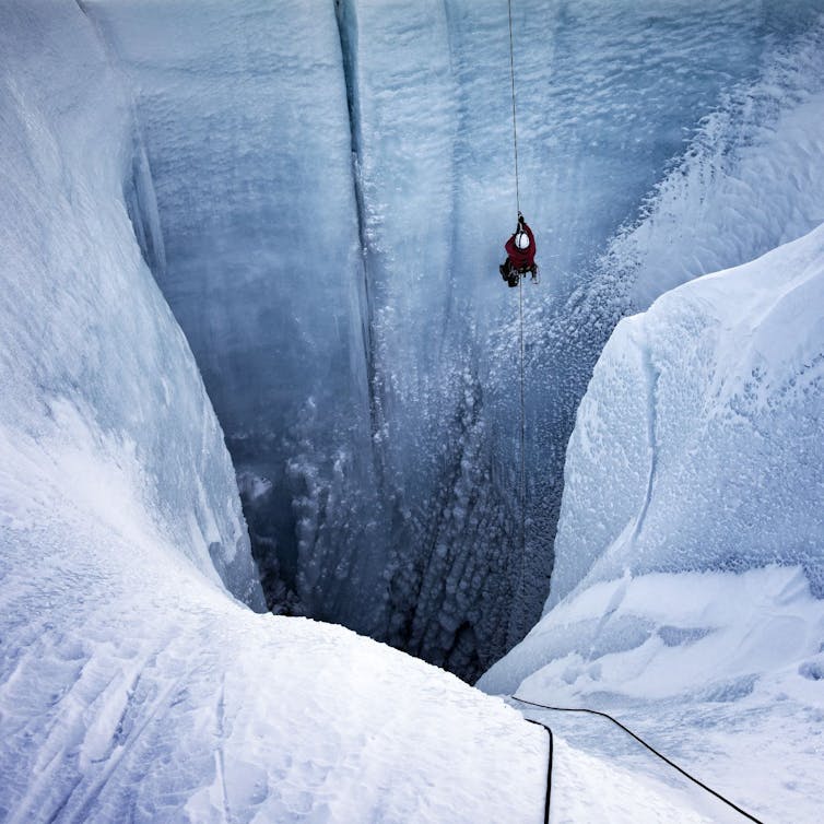 Alun Hubbard using a rappelling rope lowers himself from the top of the ice sheet into a huge hold with water pouring down the sides. The hole appears to be as wide as a two-lane road.