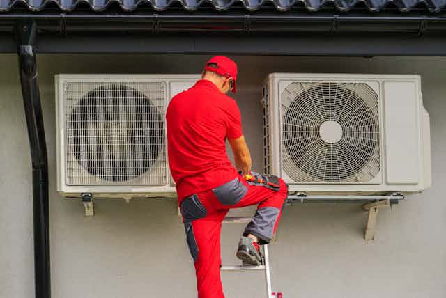 An engineer in red uniform stands atop a ladder inspecting two fan units attached to a building's exterior.