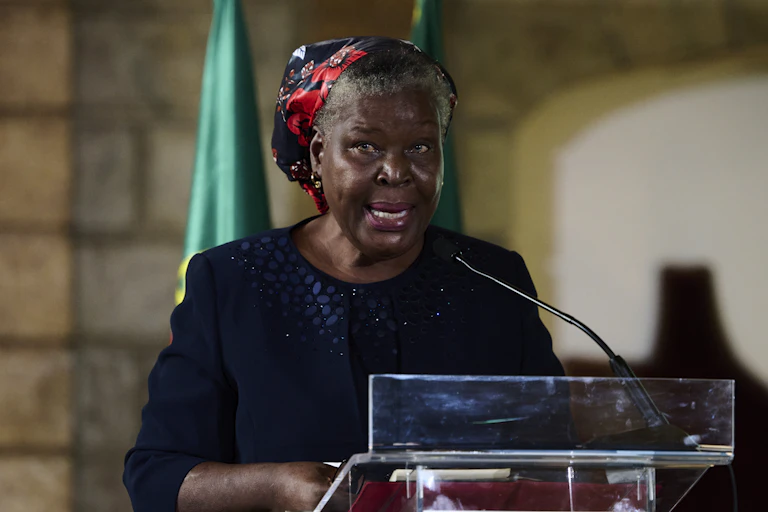 An elderly woman with greying hair and a head scarf speaks into a microphone