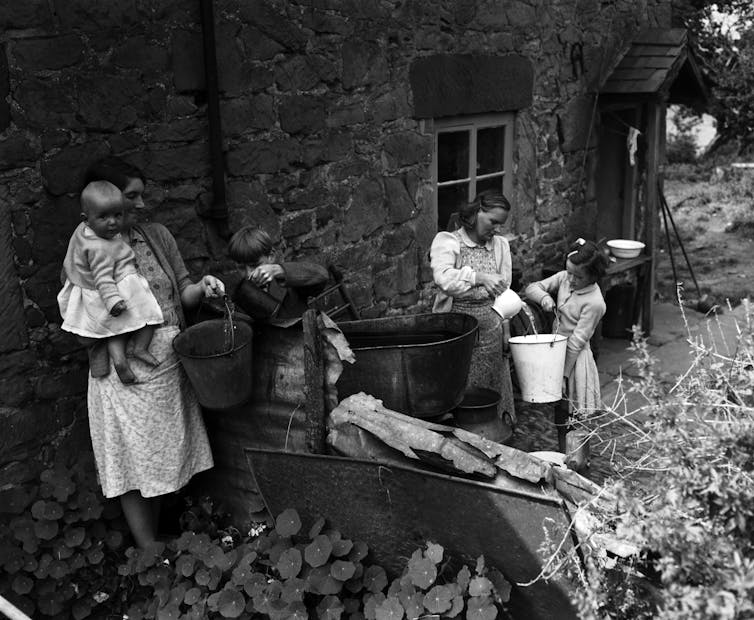 two women and three children in 1956 collect water from a tub against a stone wall of a house