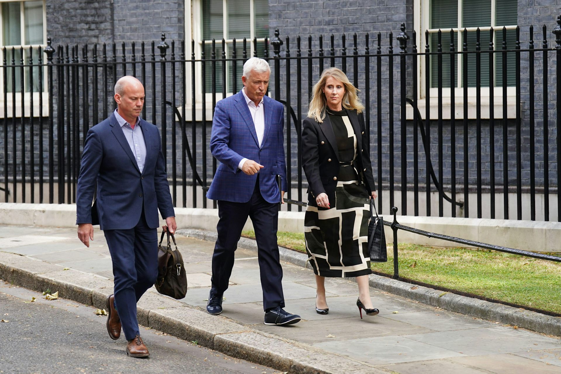 Two men in blue suits and a woman in a black and white dress and jacket walk past black railings.