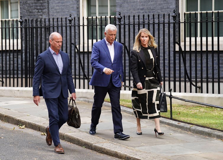 Two men in blue suits and a woman in a black and white dress and jacket walk past black railings.