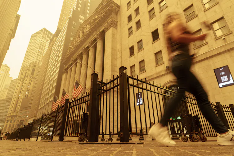 A woman walks past the New York Stock Exchange building in the Wall Street district of New York. The sky is yellow-orange with wildfire smoke, a sky color common in apocalyptic films.