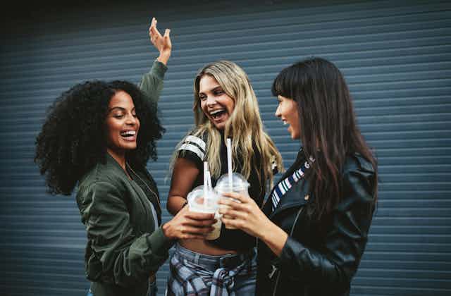 Three young female friends smiling and toasting iced coffees in front of a garage door