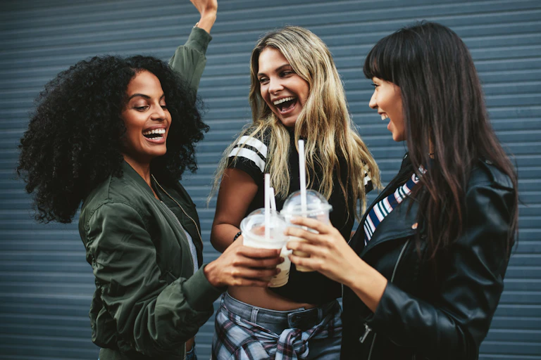Three young female friends smiling and toasting iced coffees in front of a garage door