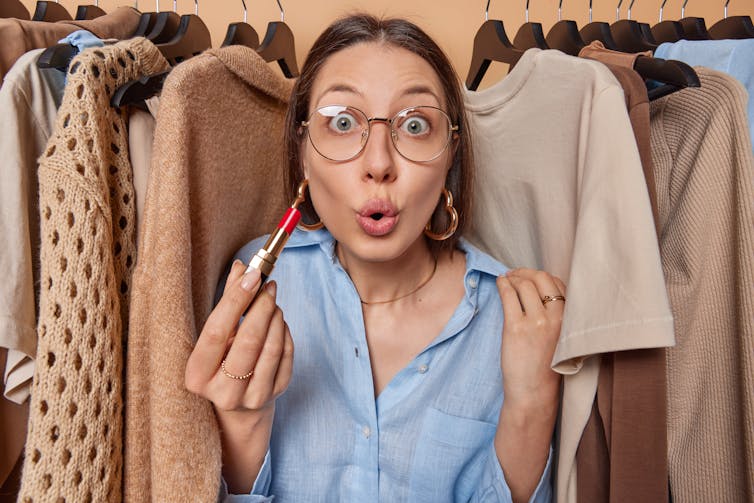 A woman holding a lipstick among a clothing rack of beige clothes