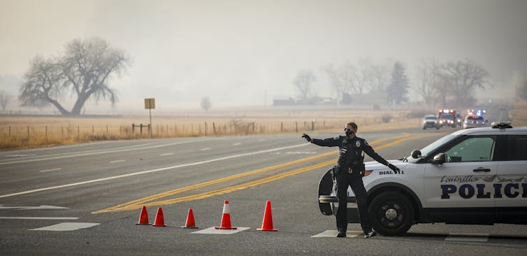 A police officer stands by traffic cones waving cars away from a smoky road.