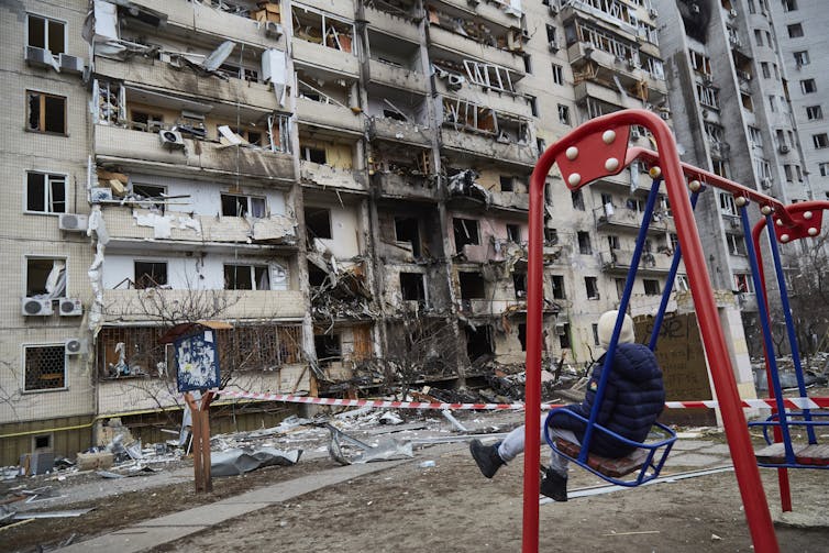 A child swings in front of a destroyed, burned looking building.