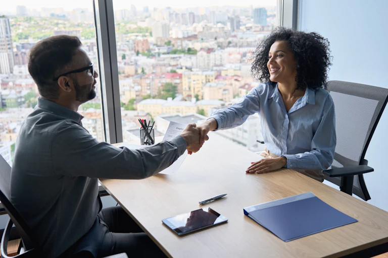 A young man shakes the hand of a woman at a desk by a large window