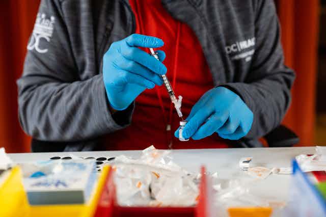 Someone wearing a red shirt, gray sweatshirt and blue medical gloves prepares a syringe.