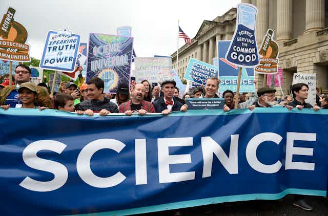 A large crowd of people hold a banner reading "Science" while others hold science with slogans like "Science is universal" and Science: protecting our communities"